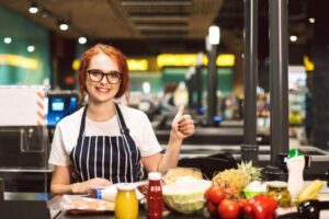 Young smiling female cashier in eyeglasses and striped apron wit