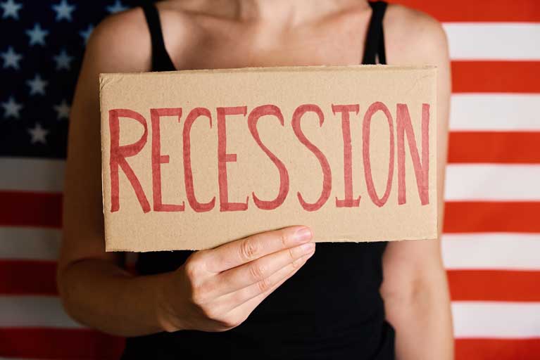 Woman standing in front of a united states flag holding up a cardboard sign with the word recession written on it.