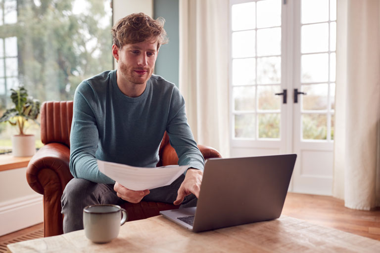 Man sitting at home checking paperwork while typing on his laptop