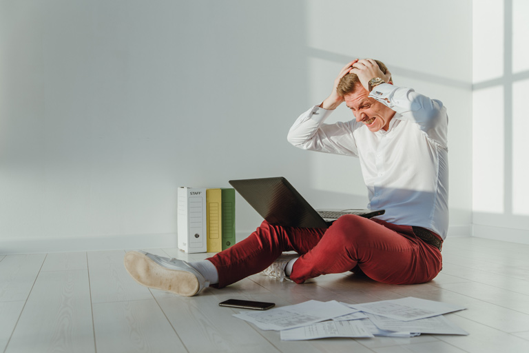 Frustrated young businessman holding his head while looking at his laptop with paperwork on the floor