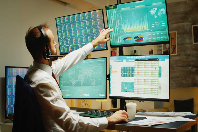 Stock market analyst looking at different stocks on 4 computer screens while drinking coffee