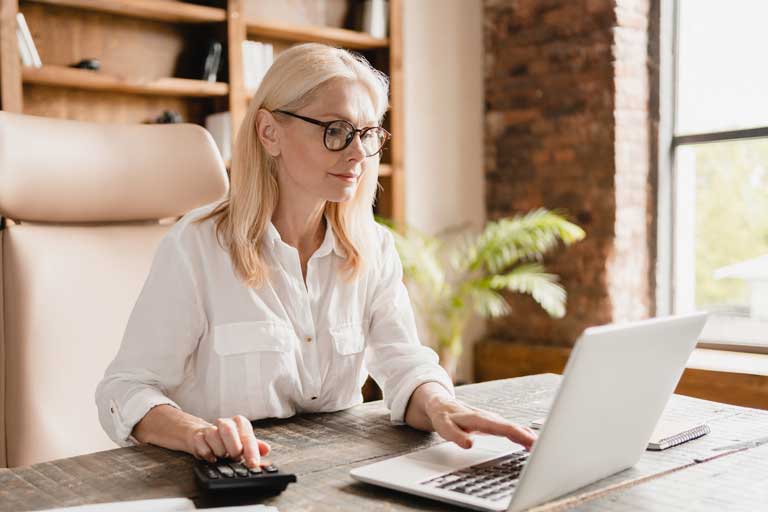 A woman working at her computer. She is wearing a white blouse and using a calculator.