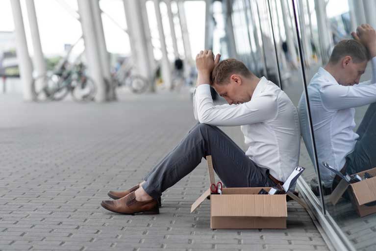 Man that just got fired from his job sitting on the floor against a building with his belongings inside a box next to him