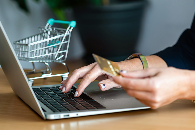 Woman with a small shopping cart next to her typing the numbers of her credit card on a laptop