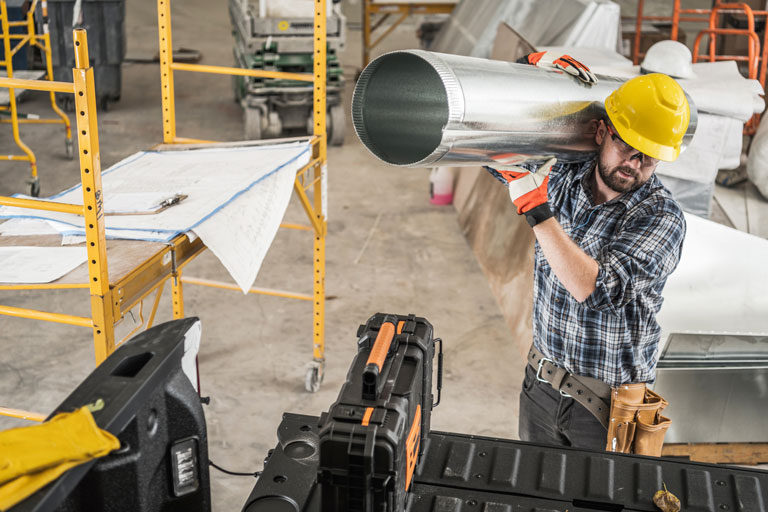 Construction worker with a yellow hard hat carrying an aluminum cylinder to the back of his pickup truck