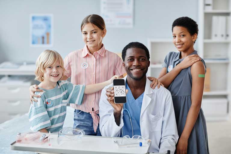 Three children wearing vaccination stickers stand around a smiling doctor who is holding up a QR code on his phone