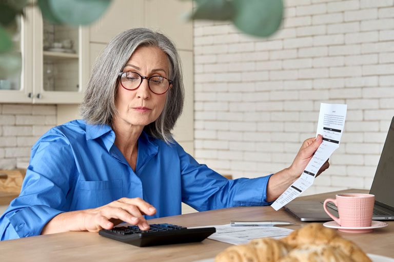Middle age woman sitting inside her kitchen typing on a calculator with several statements around her