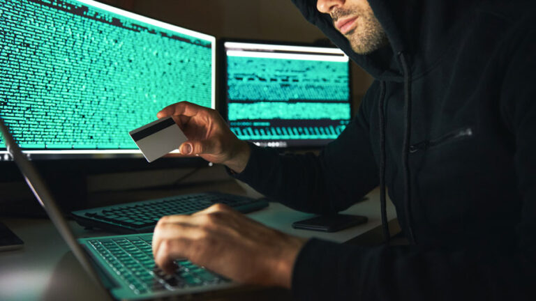 Hacker with black hoodie holding up a stolen credit card and typing on a computer