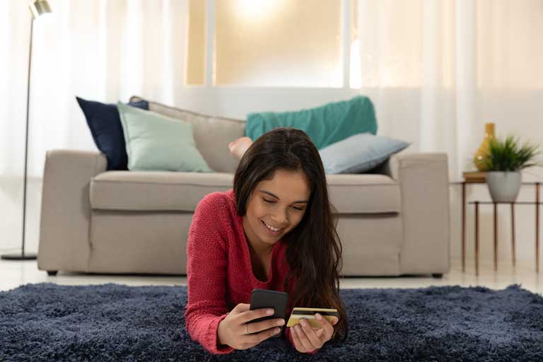 Young woman making a purchase through a smartphone while laying on the floor of her living room